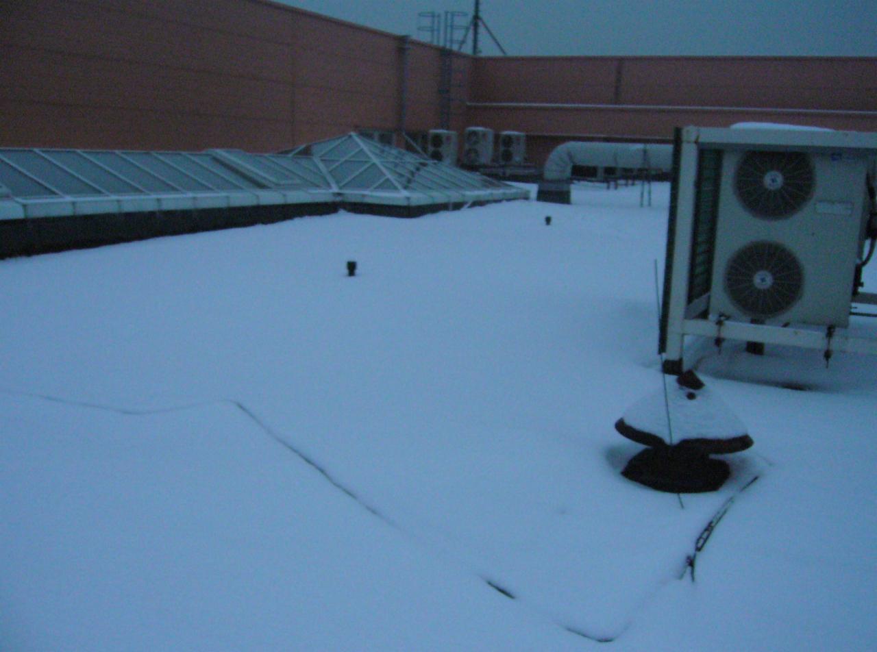 Zasypany śniegiem dach budynku z widocznymi świetlikami, wentylacją i klimatyzacją. Zimowy krajobraz miejski z elementami infrastruktury technicznej.