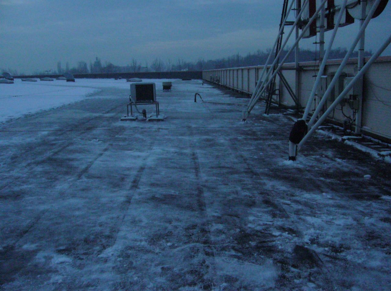 Zimowy widok na płaski dach pokryty śniegiem, z instalacjami technicznymi i odśnieżonymi śladami. Chłodna kolorystyka i pochmurne niebo.