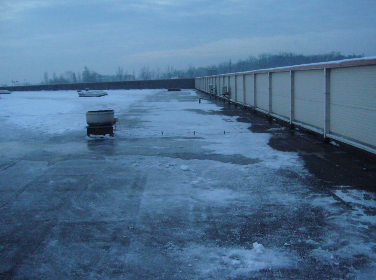 Zimowy widok na płaski dach budynku z resztkami śniegu i lodem, z widoczną instalacją wentylacyjną i ścianą z paneli w tle, pochmurne niebo.