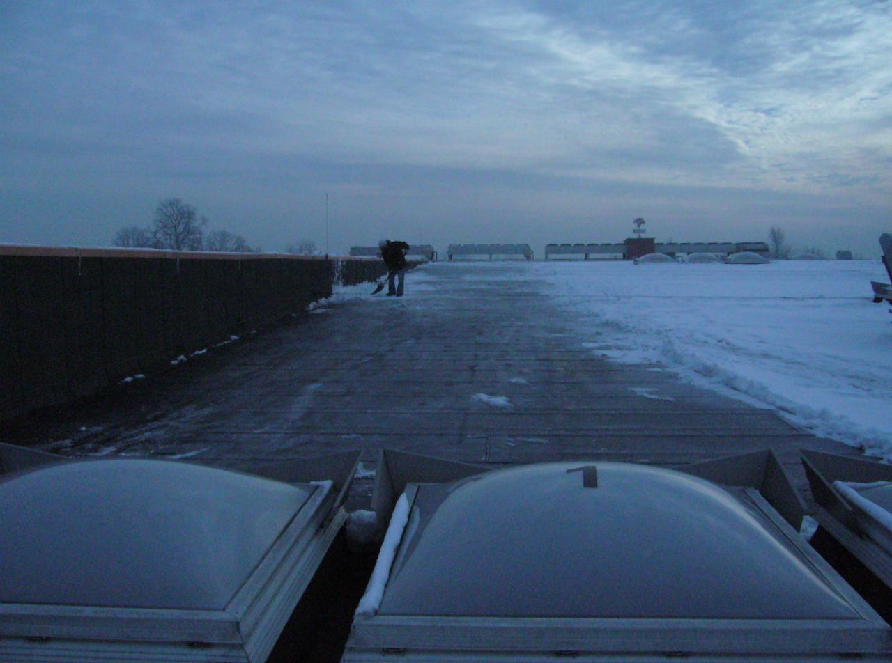 Czyszczenie dachu ze śniegu, widok z dołu na płaski dach z świetlikami, osoba odśnieża dach łopatą, pochmurne niebo w tle.