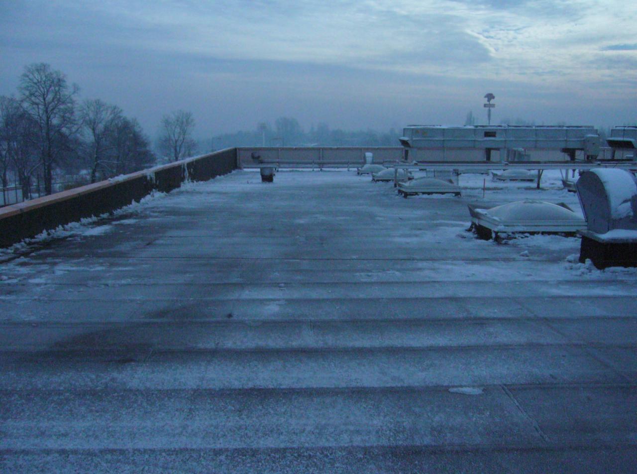 Zimowy krajobraz płaskiego dachu pokrytego śniegiem, z widocznymi elementami wentylacji i fragmentem murku. W tle drzewa i pochmurne niebo o świcie.