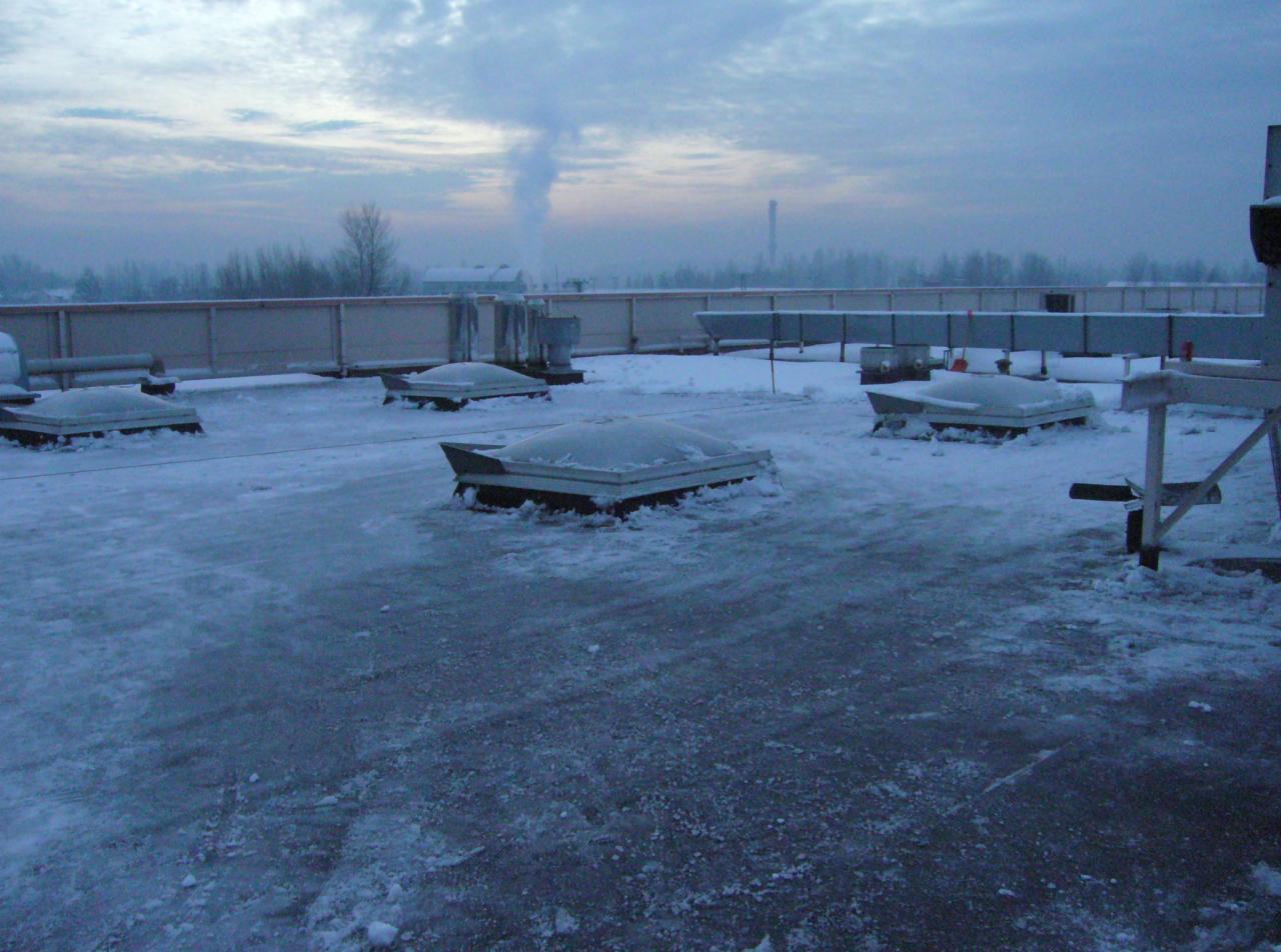 Zimowy widok na płaski dach pokryty śniegiem, z widocznymi świetlikami dachowymi i metalową barierką. W tle zarys miasta i komin.