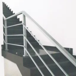 Nowoczesne schody z ciemnego kamienia z minimalistyczną, metalową balustradą. Widok z dołu, oświetlenie dzienne. W tle biała ściana i fragment grzejnika.