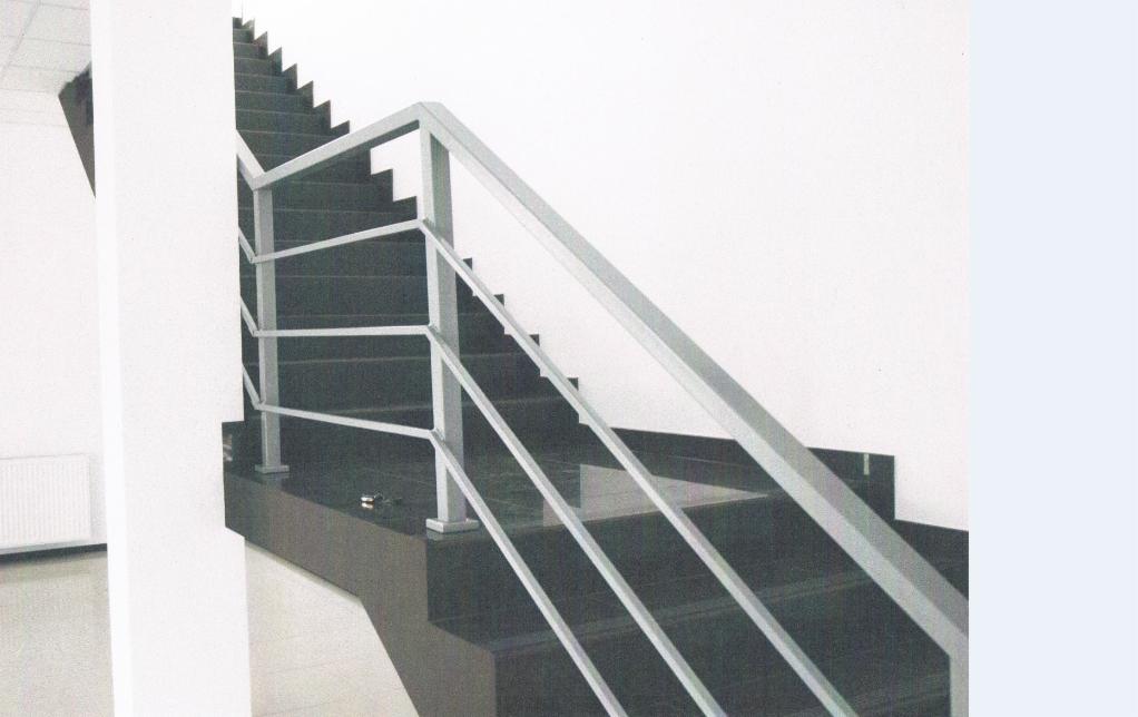 Nowoczesne schody z ciemnego kamienia z minimalistyczną, metalową balustradą. Widok z dołu, oświetlenie dzienne. W tle biała ściana i fragment grzejnika.