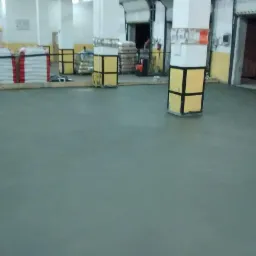 Szara, gładka posadzka w hali przemysłowej, widoczne worki z materiałami budowlanymi, żółto-czarne osłony kolumn i maszyna do polerowania posadzek w prawym dolnym rogu.