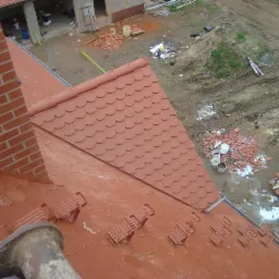 Czerwony dach z dachówki karpiówki w Kobylinie, widok z góry. Na dachu stopnie kominiarskie, w tle komin z cegły i teren budowy.