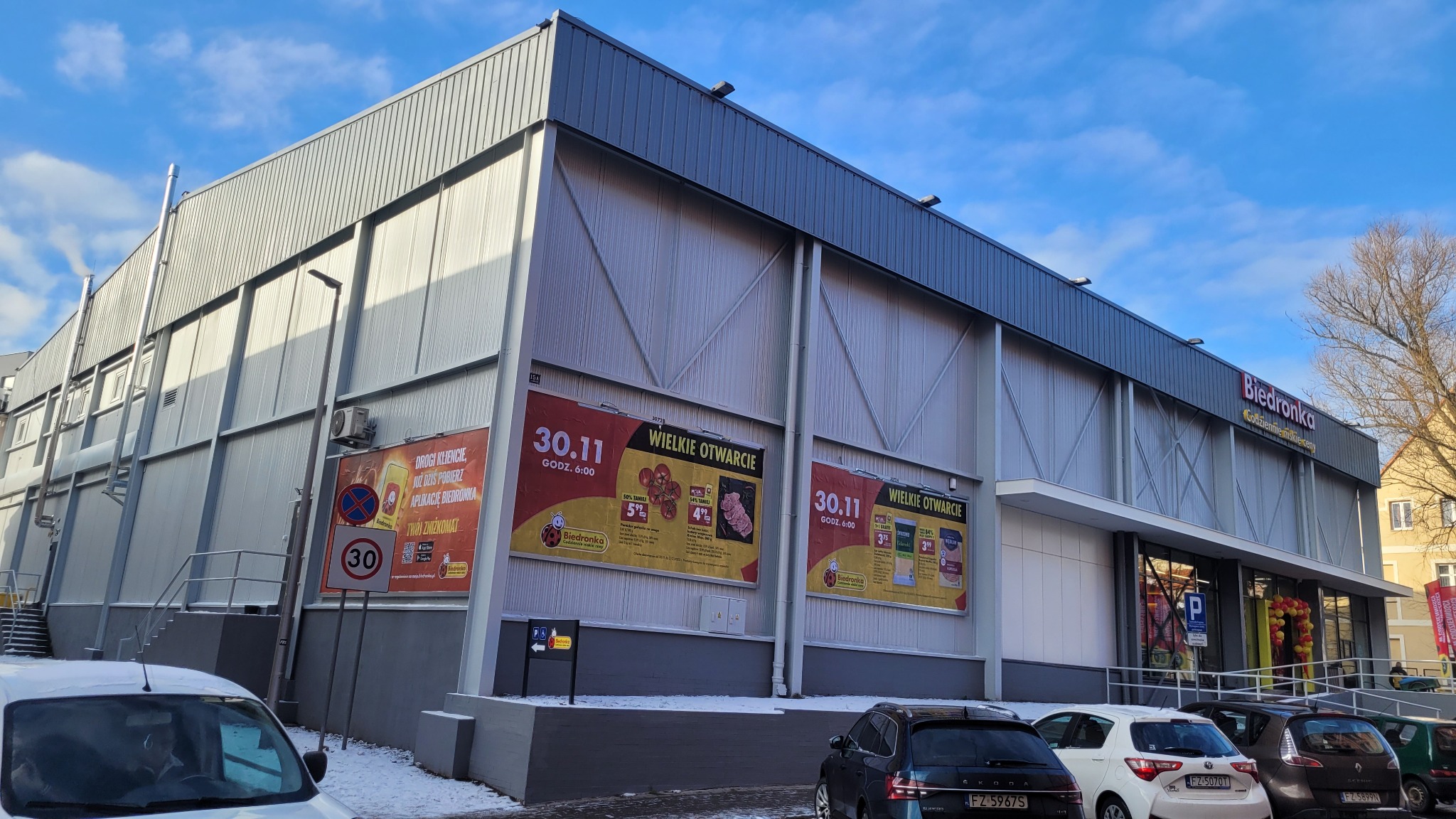 Nowoczesny budynek sklepu wielkopowierzchniowego z metalową elewacją w kolorze szarym, z dużymi plakatami reklamowymi 'Wielkie Otwarcie' i widocznymi zaparkowanymi samochodami na pierwszym planie.