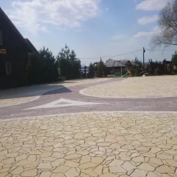 Beżowa kostka brukowa ułożona w wz&oacute;r na placu przed Karczmą Rohatyną. Ciemniejsze pasy kostki tworzą ścieżkę i skrzyżowanie.