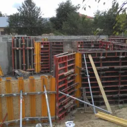 Przygotowanie szalunków i zbrojenia pod wylewanie fundamentów domu jednorodzinnego na działce z widokiem na budynek gospodarczy i drzewa.