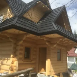 Wejście do domu z bali z czarnym dachem, widoczne detale konstrukcyjne i sąsiedni budynek z czerwonym dachem w tle.