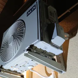 Zewnętrzna jednostka klimatyzatora marki AUX zamontowana na elewacji budynku z drewnianym podbitką dachową, widoczne białe rurki przyłączeniowe.