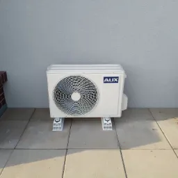 Biały klimatyzator AUX na zewnątrz, na betonowej posadzce, z widocznym fragmentem ceglanej ściany i niebieską donicą z kwiatami. Szara ściana w tle.