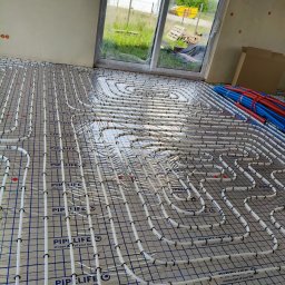 EL-BUD Robert Makuch - Ogrzewanie podłogowe w Owińskach: białe rurki instalacji ułożone na folii z siatką, widoczne niebieskie i czerwone rury. Okno z widokiem na zewnątrz.