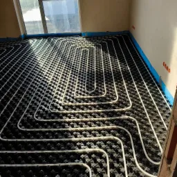 Białe rury ogrzewania podłogowego ułożone na czarnej macie systemowej, widoczne okno i fragment ściany. Instalacja w promieniach słońca.