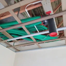 Zielone, karbowane przewody wentylacyjne ułożone pod drewnianą konstrukcją sufitu podwieszanego, widoczny element systemu z czarną rurą i czerwonymi elementami.