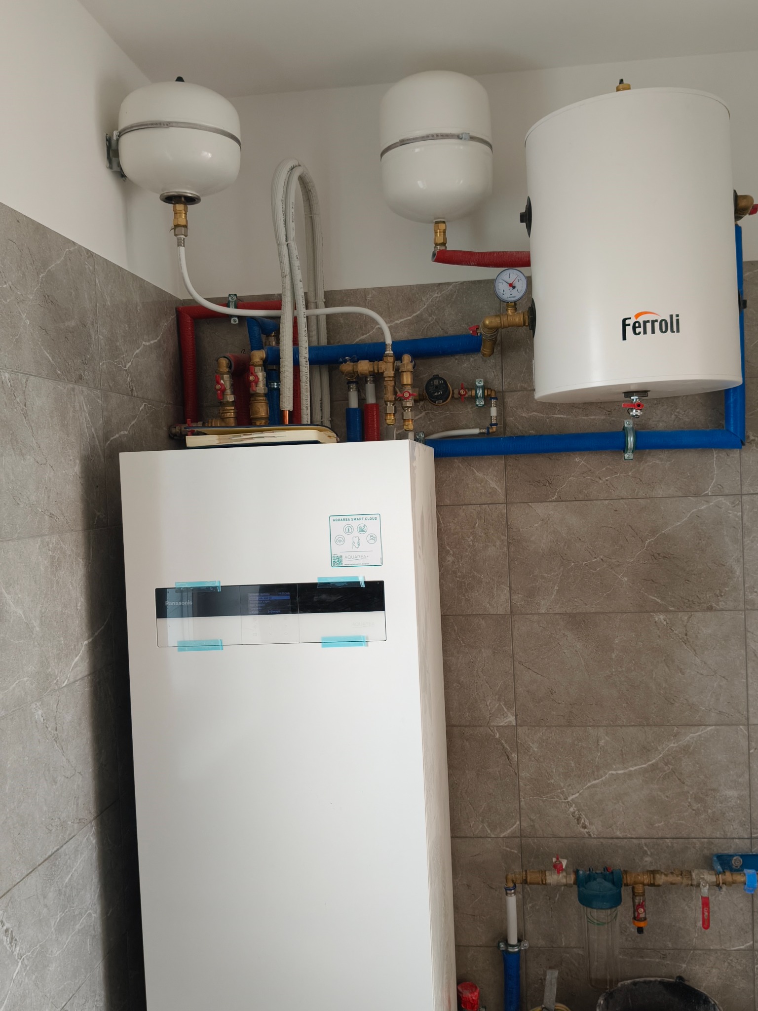 Biała pompa ciepła Panasonic Aquarea Smart Cloud z widocznymi przyłączami rur miedzianych i plastikowych, zbiornikiem Ferroli oraz dwoma zbiornikami wyrównawczymi zamontowana na tle płytek...