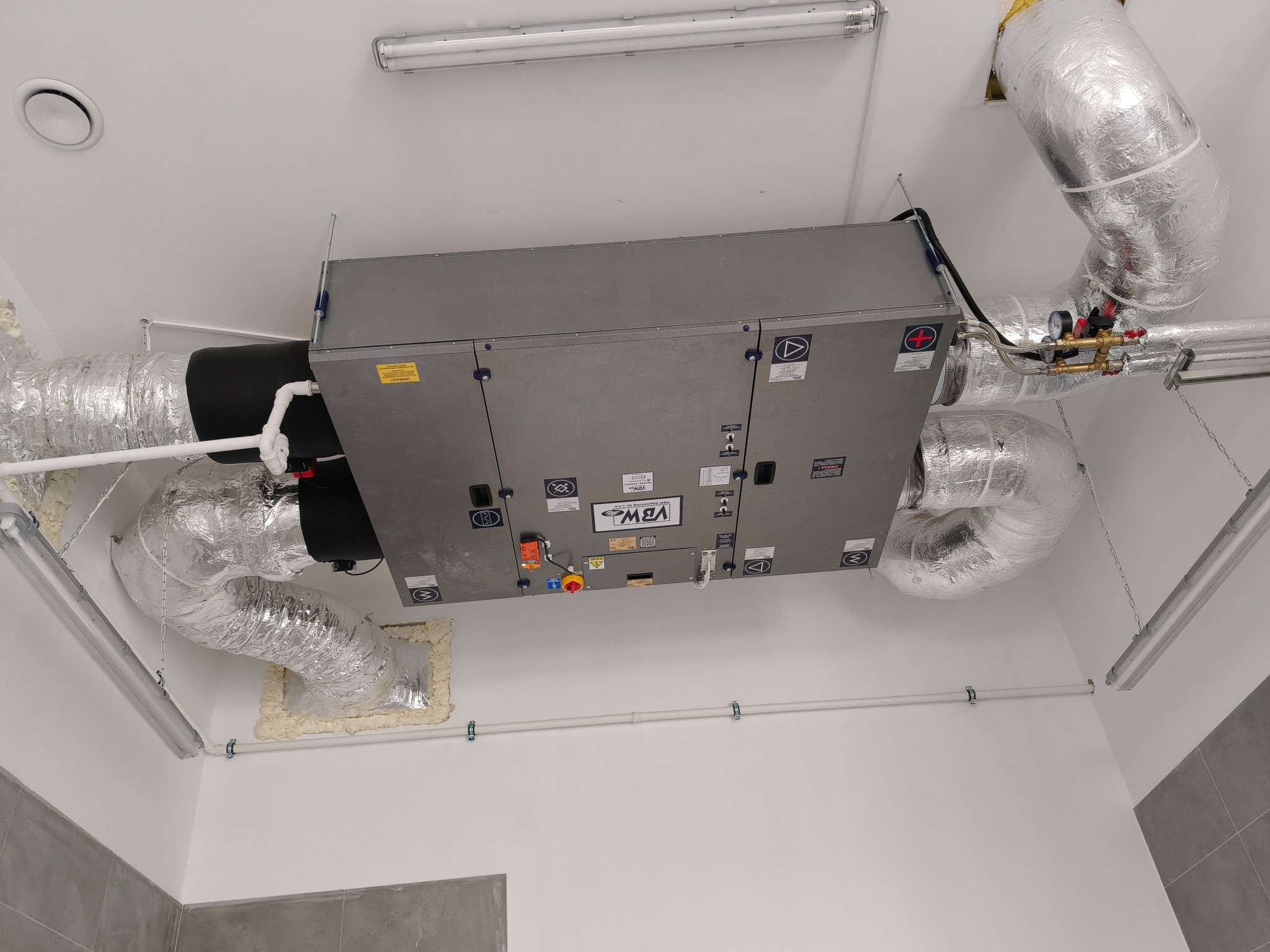 Centrala wentylacyjna z rekuperacją, widok od dołu, z widocznymi przewodami wentylacyjnymi owiniętymi folią aluminiową i elementami mocującymi do sufitu.