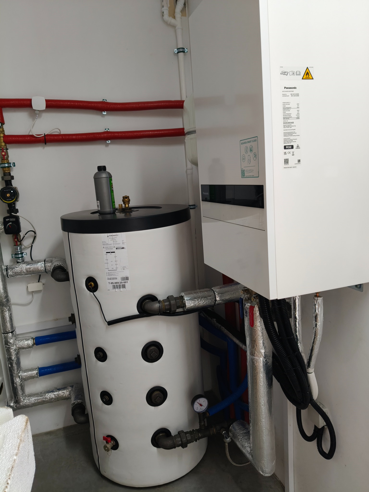 Biały zasobnik ciepłej wody z czarnym blatem, obok pompa ciepła Panasonic, widoczne rury w izolacji aluminiowej i czerwonej, oraz butelka oleju na zbiorniku.