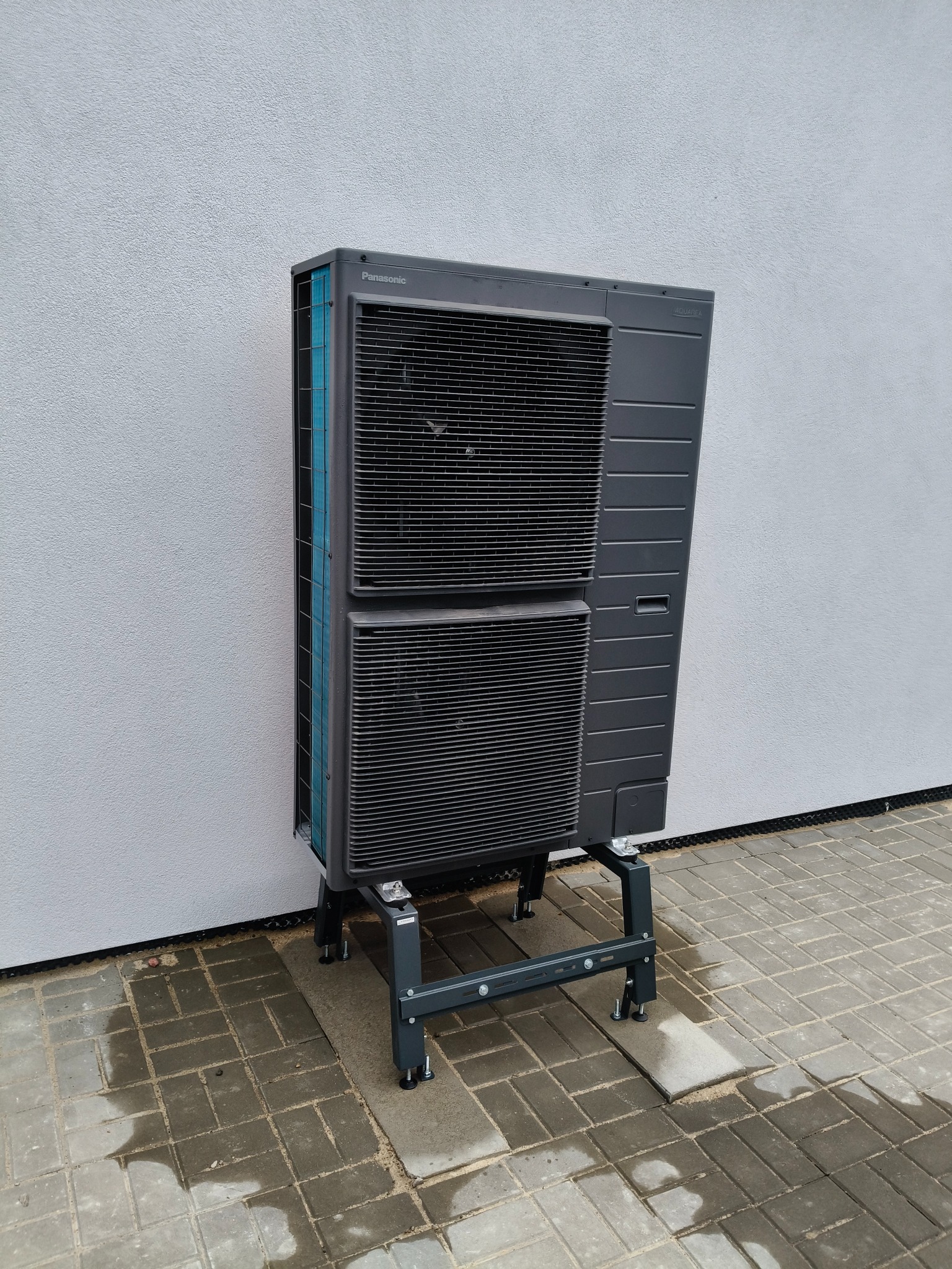 Zewnętrzna jednostka pompy ciepła Panasonic Aquarea na metalowej podstawie z regulowanymi nóżkami, ustawiona na szarej kostce brukowej przy jasnej ścianie budynku.