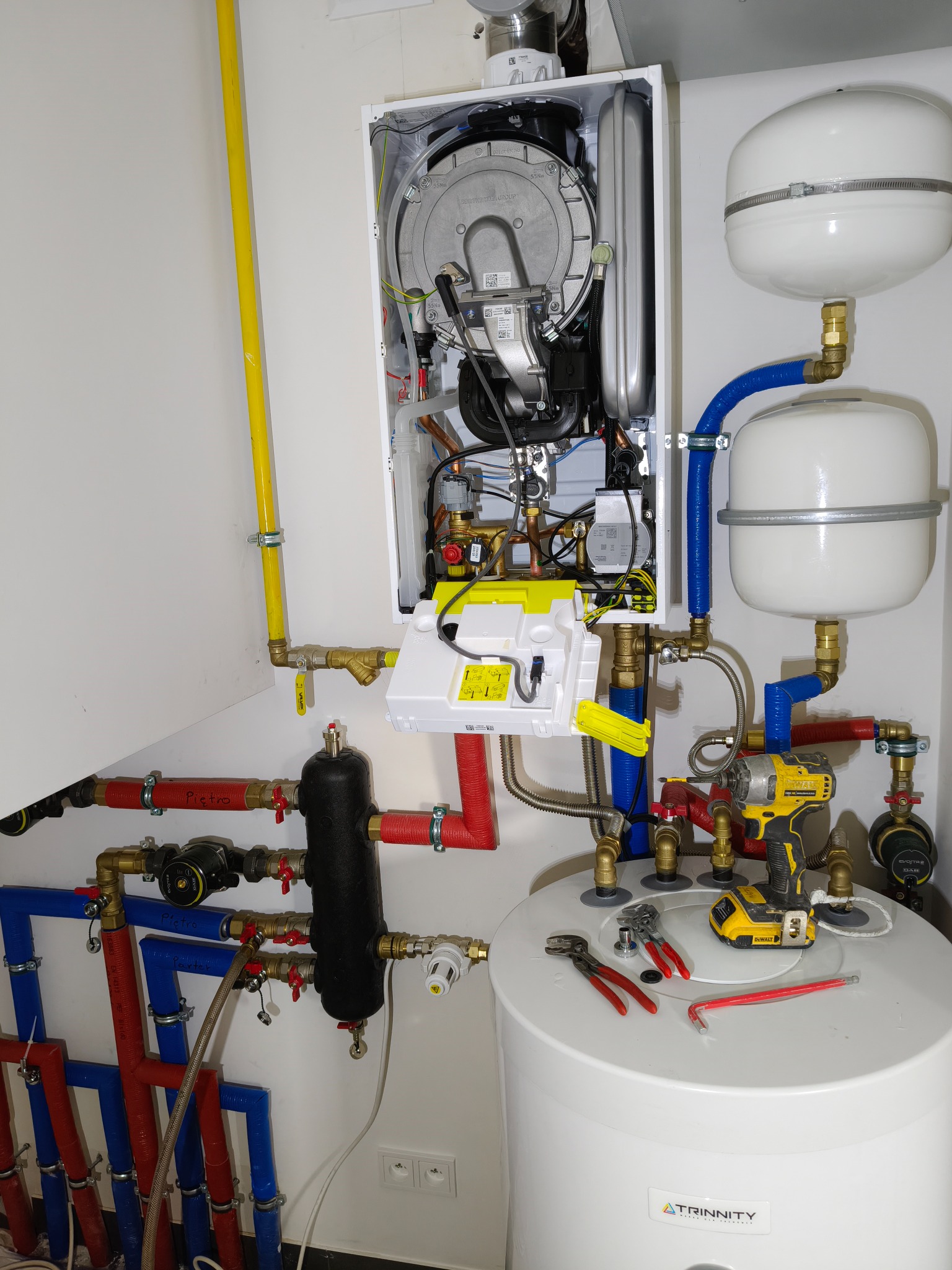 Instalacja kotła grzewczego z podłączonymi rurami i zbiornikiem, narzędzia na zbiorniku, żółta rura pionowo wzdłuż ściany.