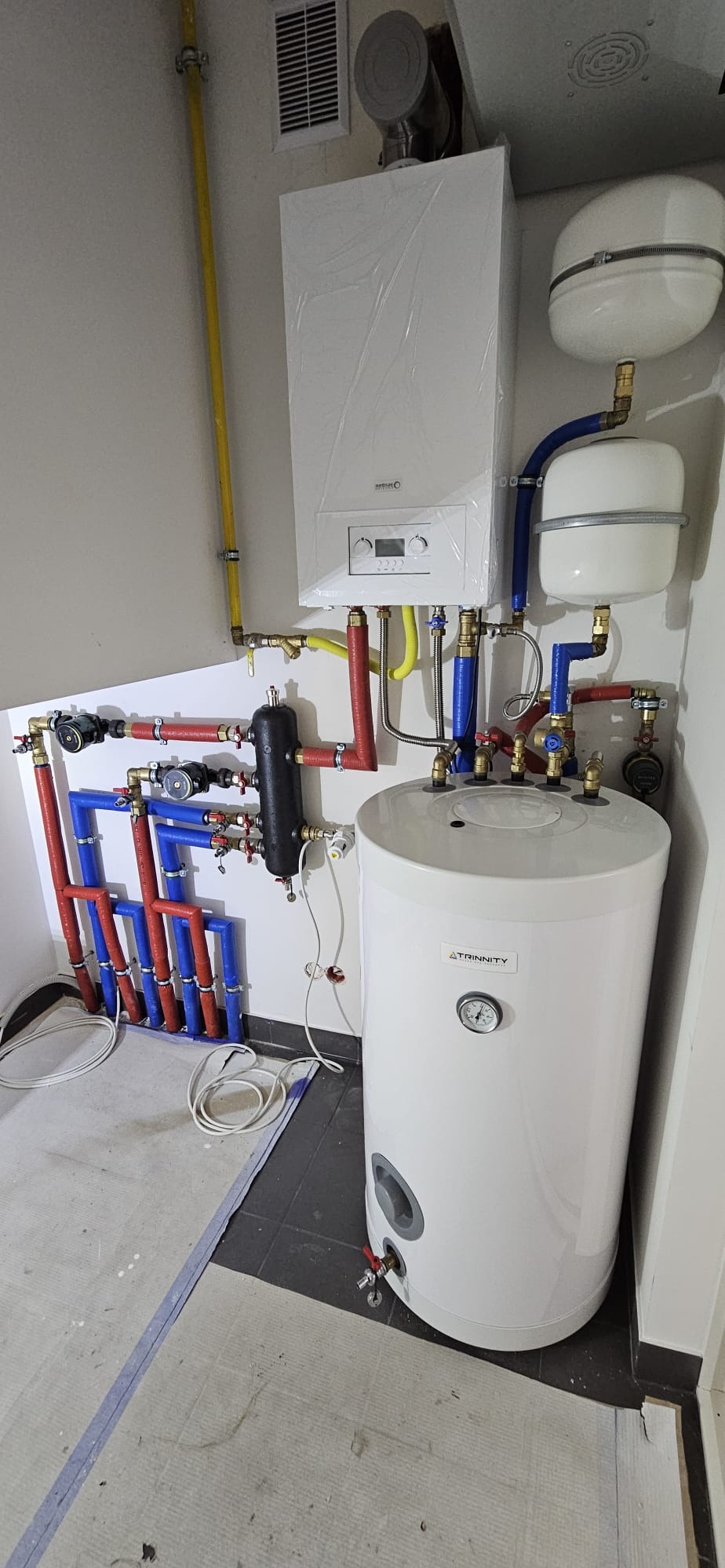 Zamontowany, biały bojler Trinity z podłączonymi rurami (czerwone, niebieskie, żółte) i osprzętem w pomieszczeniu. Na ścianie piec grzewczy w folii ochronnej.