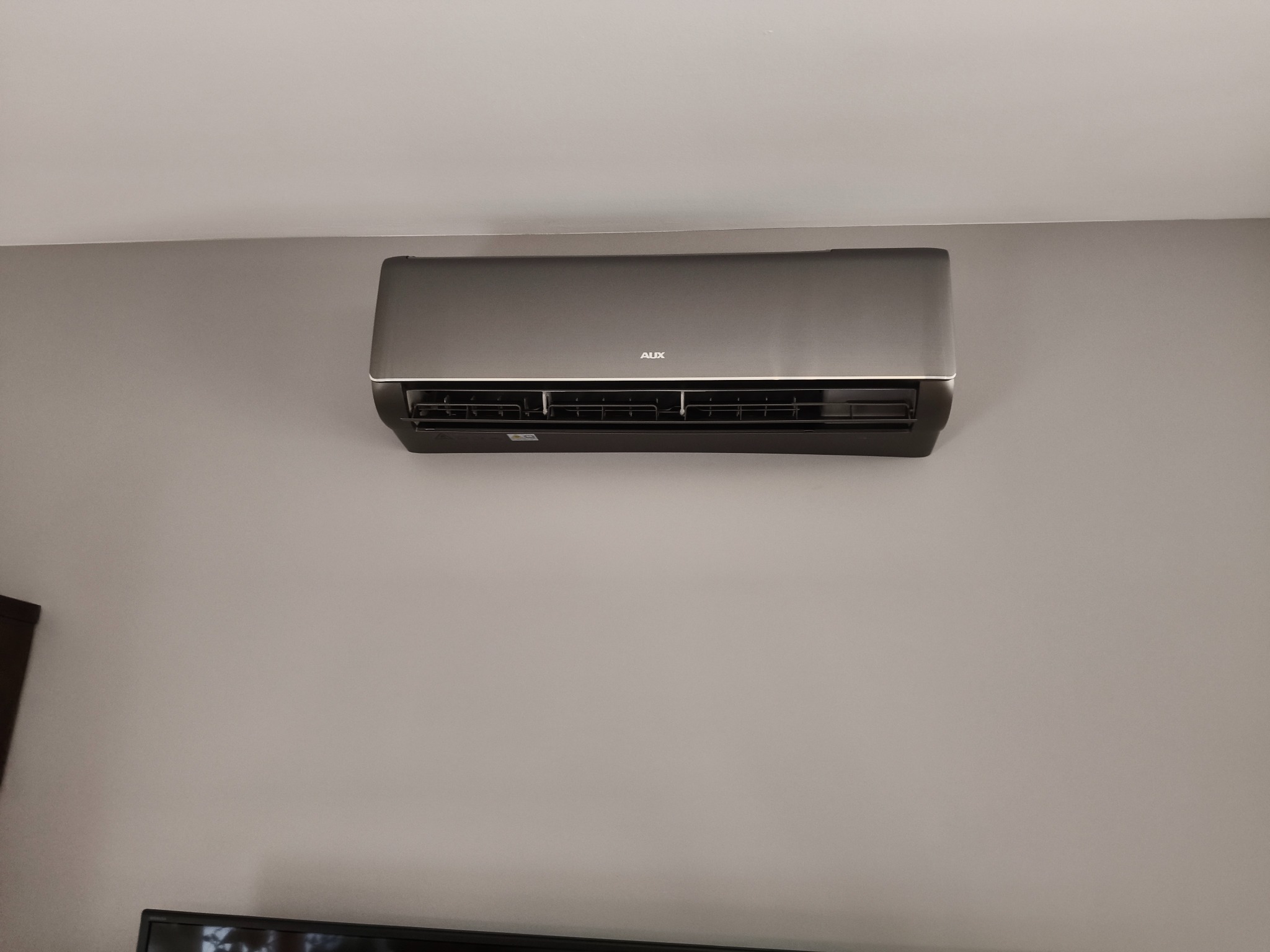 Zamontowana na szarej ścianie klimatyzacja AUX o srebrnej obudowie. Widoczne żaluzje wlotu powietrza i fragment czarnego telewizora w dolnej części kadru.