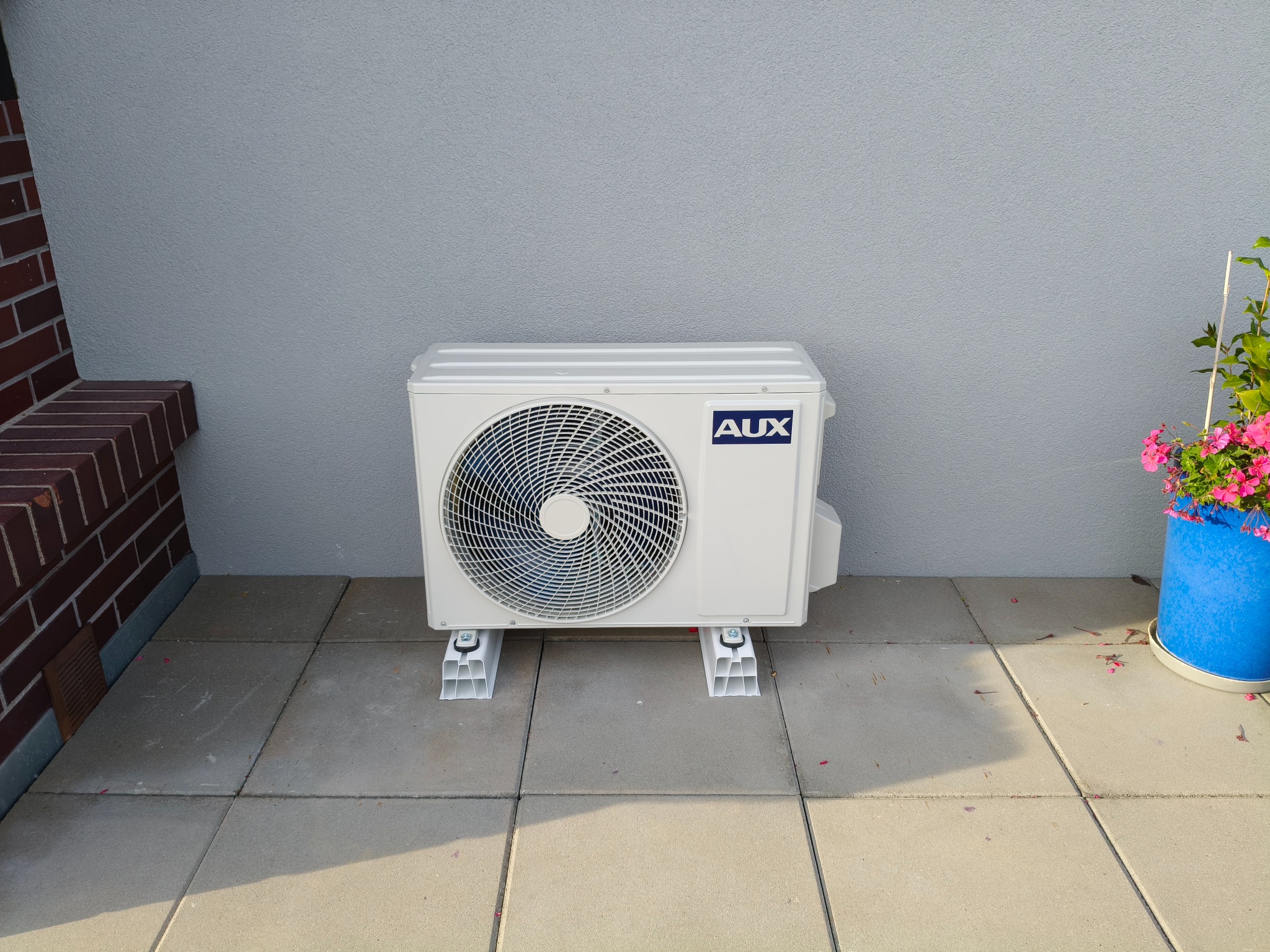 Biały klimatyzator AUX na zewnątrz, na betonowej posadzce, z widocznym fragmentem ceglanej ściany i niebieską donicą z kwiatami. Szara ściana w tle.