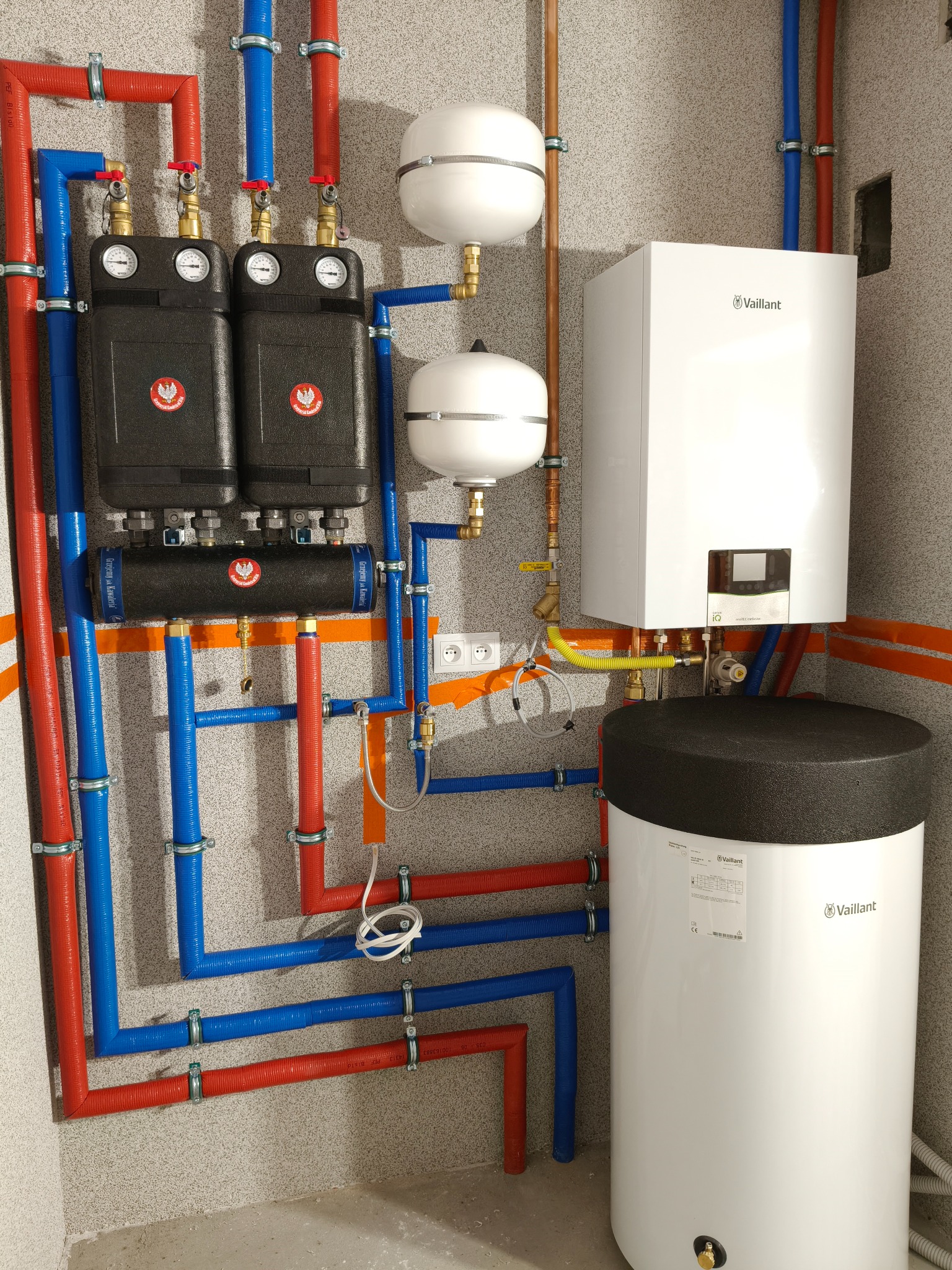 Kompletna instalacja grzewcza z piecem Vaillant, zbiornikiem i rozdzielaczem. Rury miedziane i PEX w kolorach niebieskim i czerwonym, zamontowane na szarej ścianie.