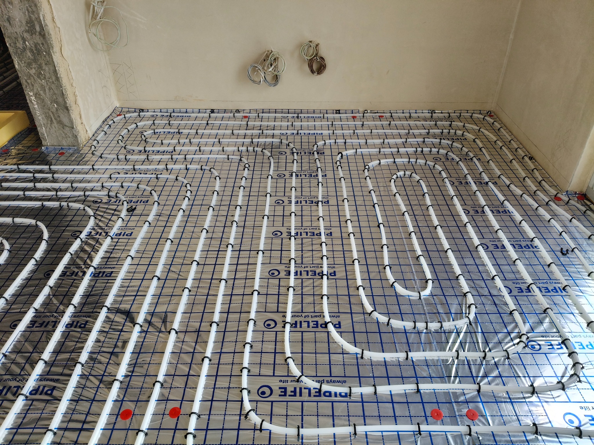 Białe rury ogrzewania podłogowego PIPELIFE rozłożone na folii z rastrem, przymocowane czarnymi uchwytami. Widoczne kable na suficie i fragment ściany w surowym stanie.