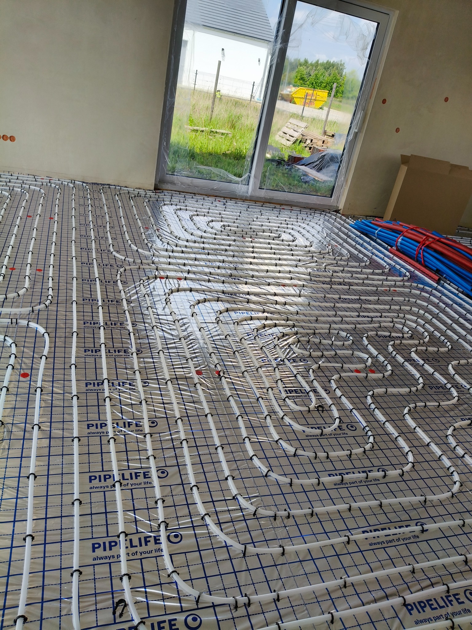 Ogrzewanie podłogowe w Owińskach: białe rurki instalacji ułożone na folii z siatką, widoczne niebieskie i czerwone rury. Okno z widokiem na zewnątrz.