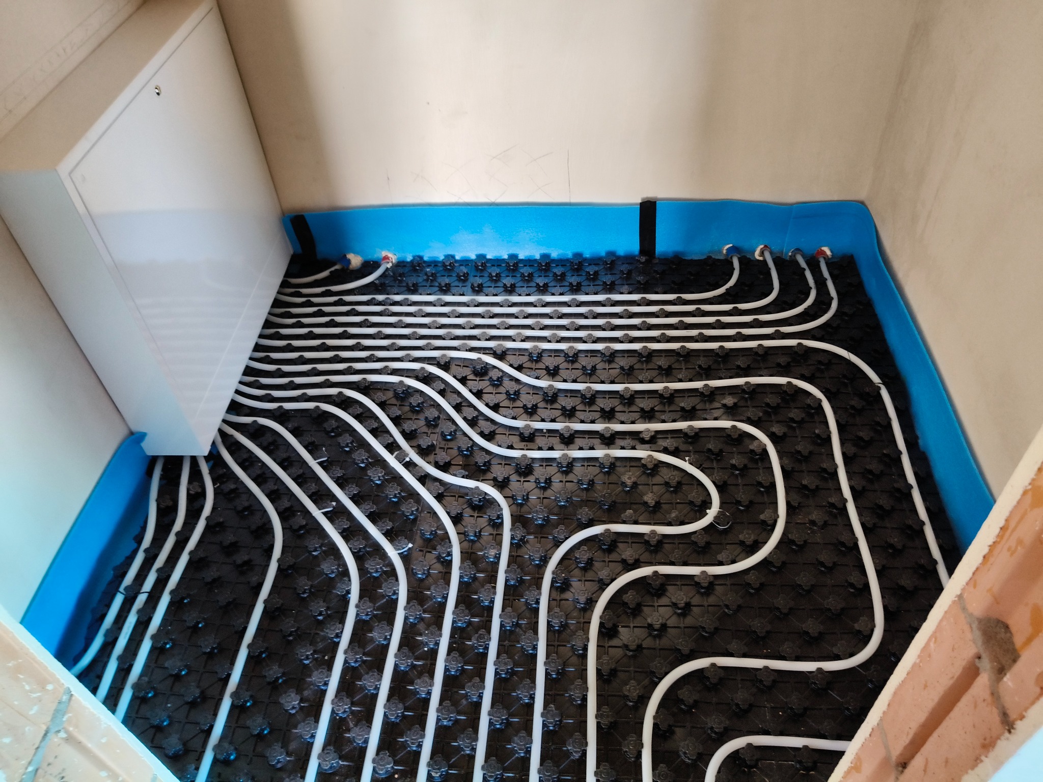 Białe rury ogrzewania podłogowego ułożone na czarnej macie z niebieską izolacją przy ścianach, widok z góry, surowy stan wykończenia.