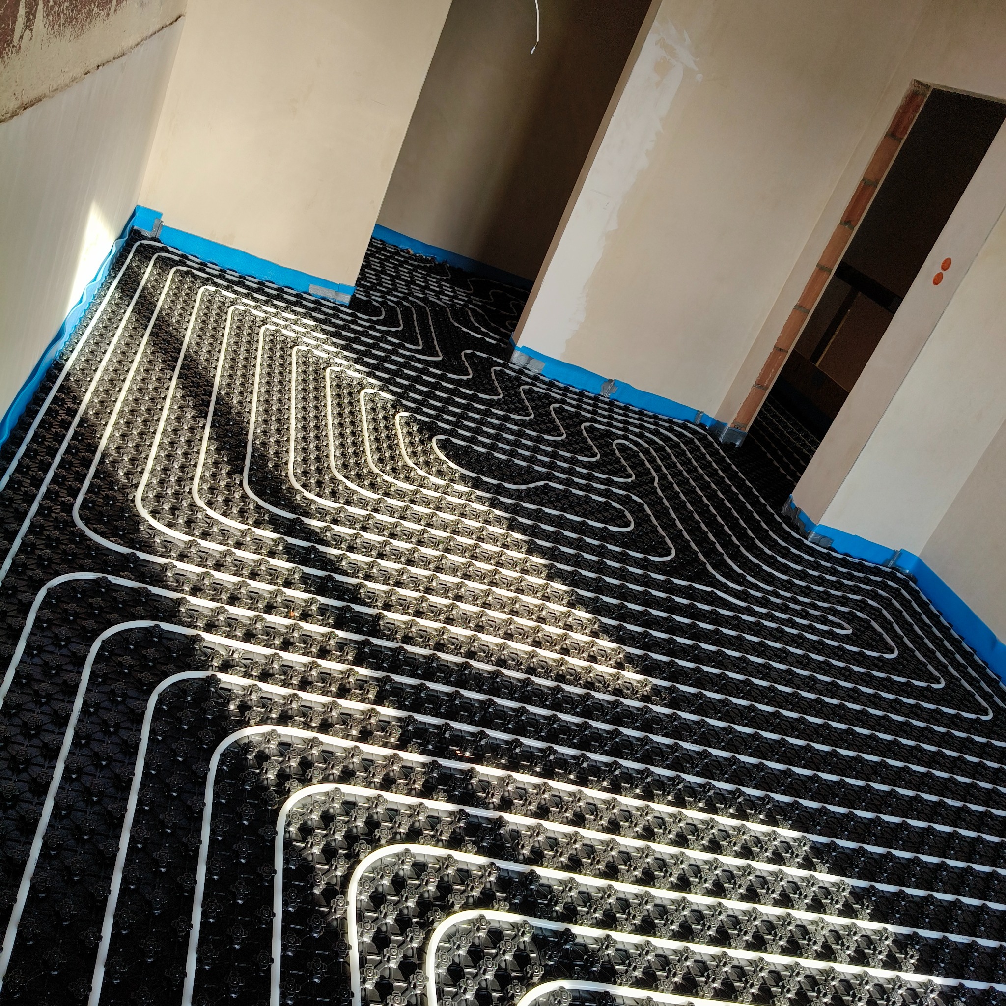 Ułożone białe rury ogrzewania podłogowego na czarnej macie izolacyjnej w surowym wnętrzu. Niebieska taśma na ścianach. Widok z góry, ukośny kadr.