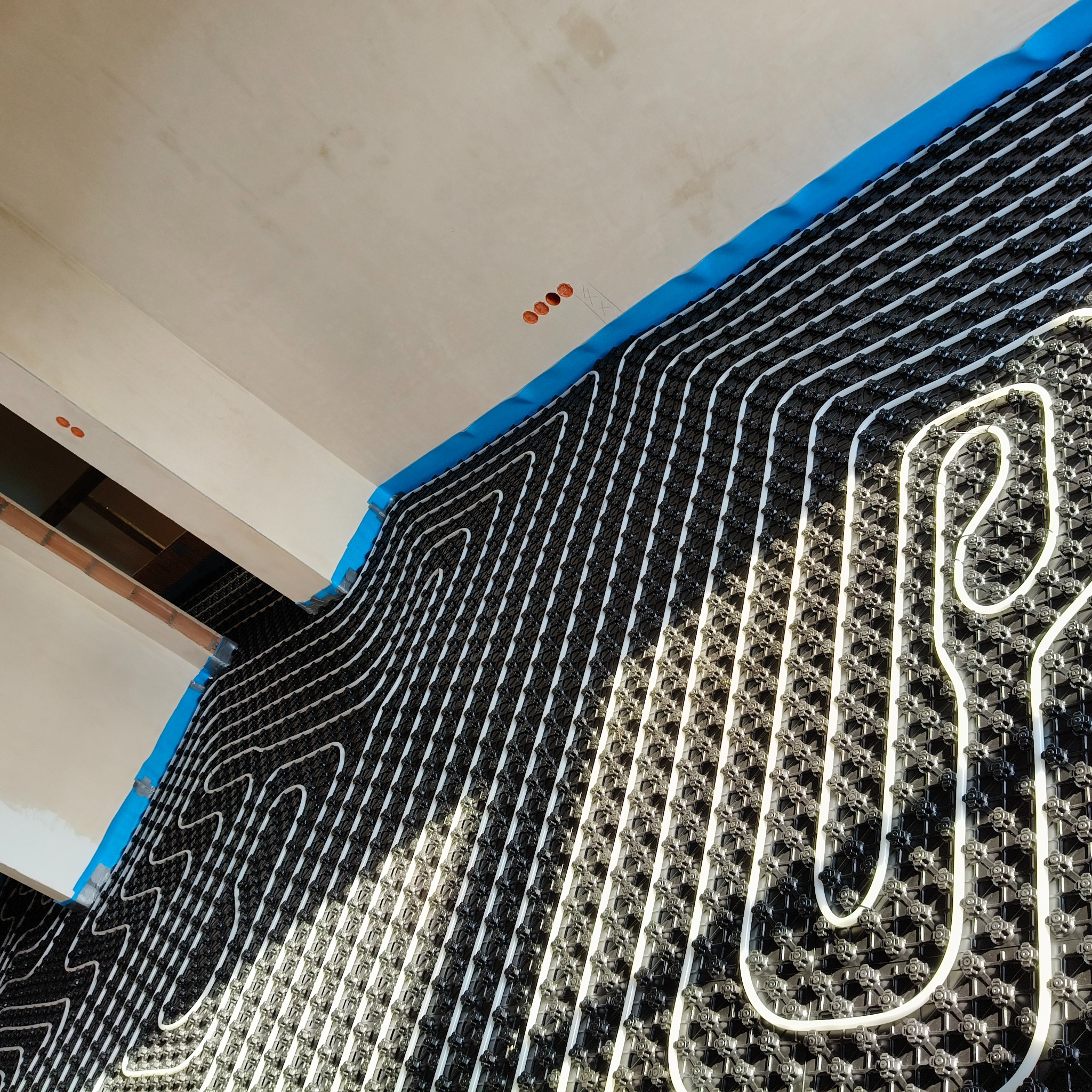 Ogrzewanie podłogowe: białe rurki wpięte w czarną matę montażową, przytwierdzone do ściany niebieską taśmą, widok z dołu w surowym wnętrzu.