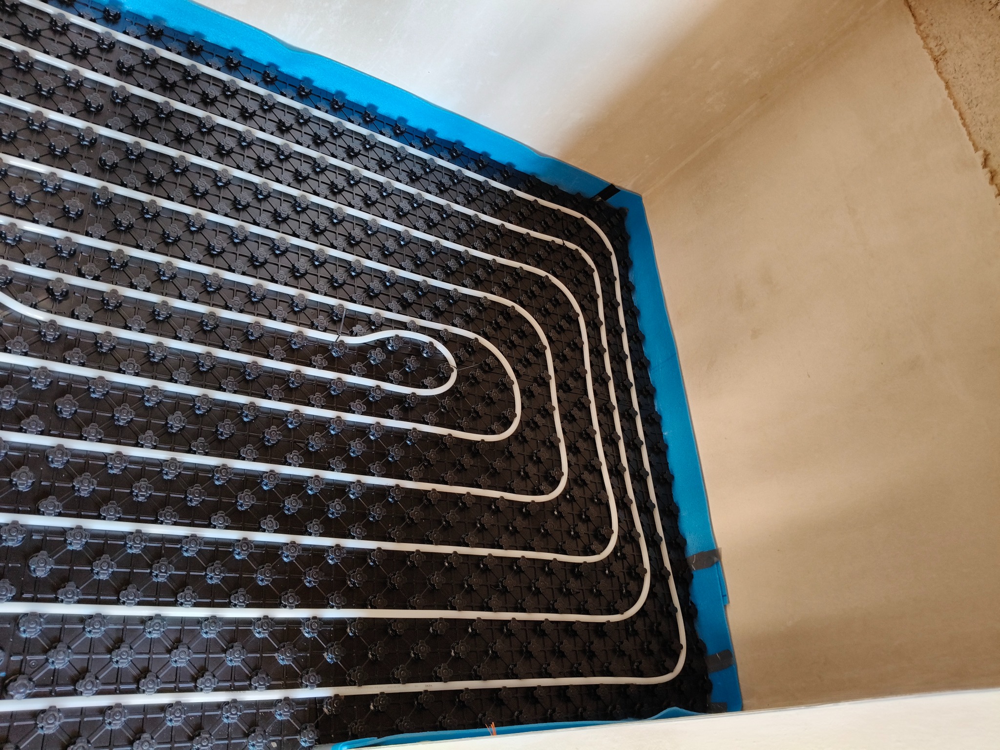 Białe rury ogrzewania podłogowego ułożone na czarnej macie z niebieską izolacją brzegową, widok z góry, surowy stan ścian.