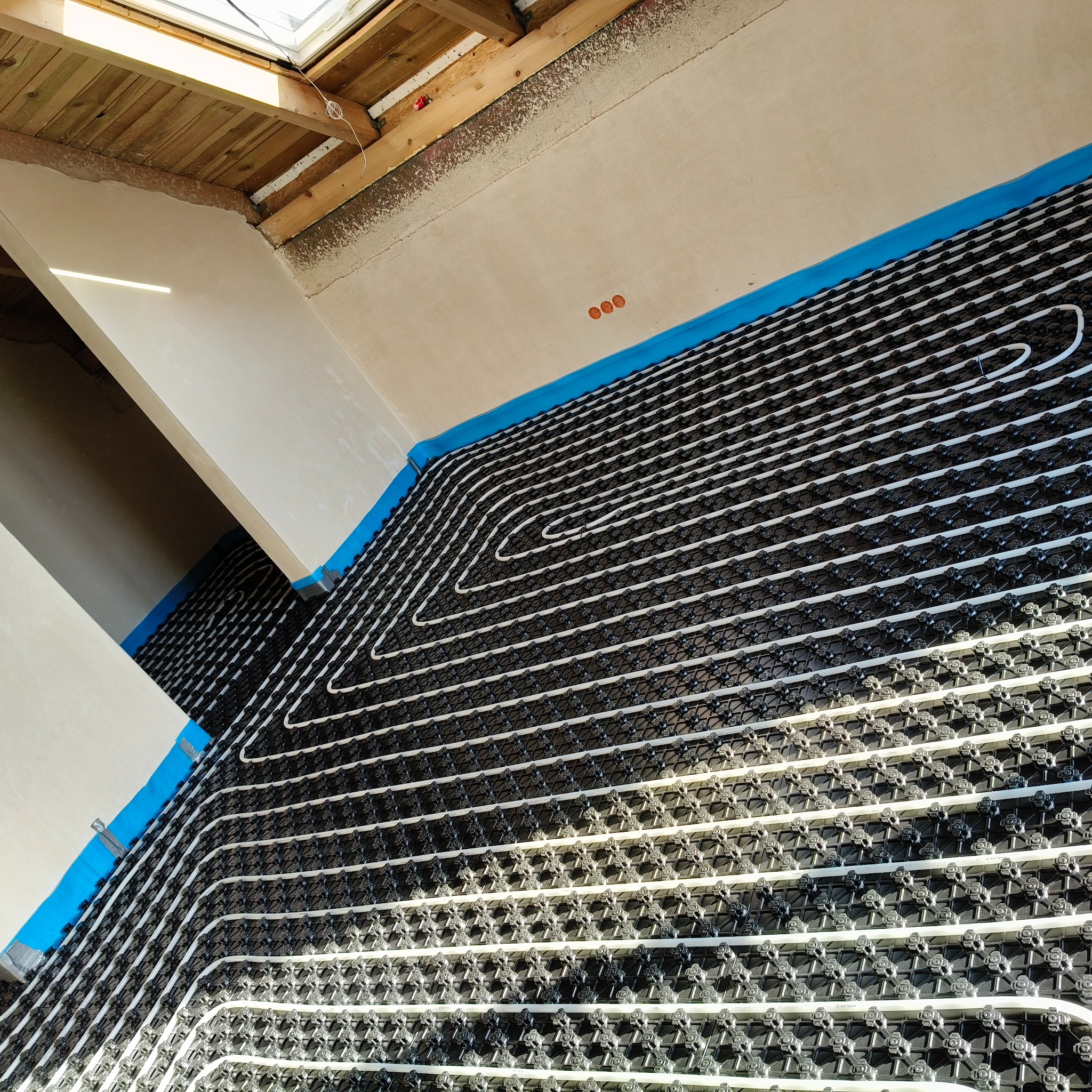 Białe rury ogrzewania podłogowego ułożone na czarnej macie w jasnym pomieszczeniu z oknem dachowym. Widoczne niebieskie taśmy mocujące rury do ścian.