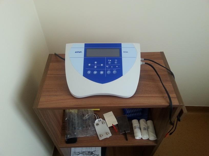 Aparat Astar Aries do elektroterapii na drewnianym regale z akcesoriami, widok z góry, kabel zasilający po prawej stronie.