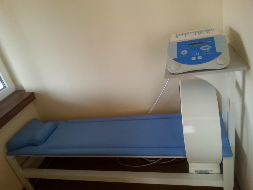 Niebieski stół do terapii z aparaturą, widok z boku. Sprzęt medyczny w gabinecie rehabilitacyjnym, jasne ściany i okno. Urządzenie do fizykoterapii.