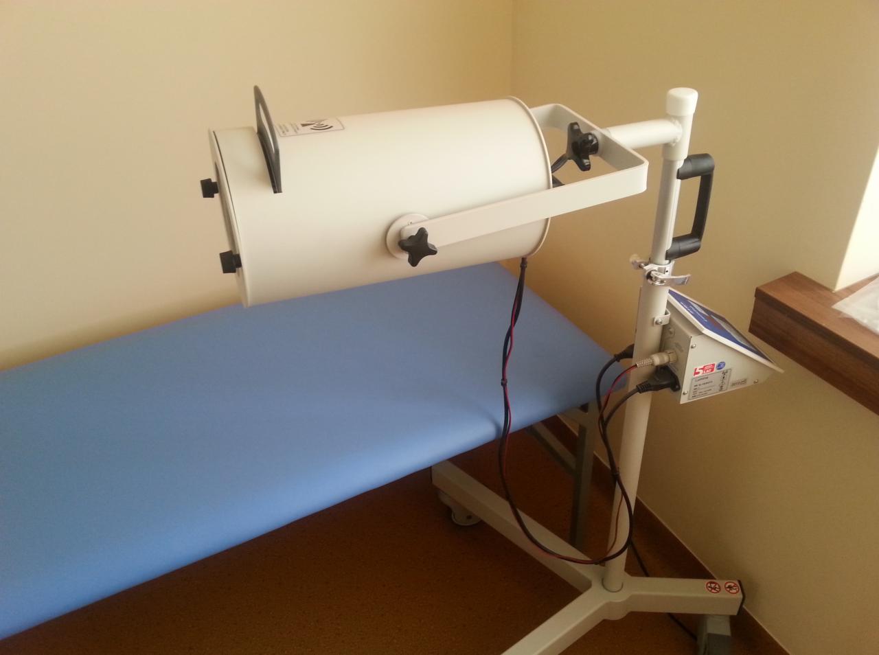 Lampa do naświetleń na statywie obok niebieskiego leżaka w gabinecie. Sprzęt do fizykoterapii z widocznymi kablami i regulowanymi elementami.
