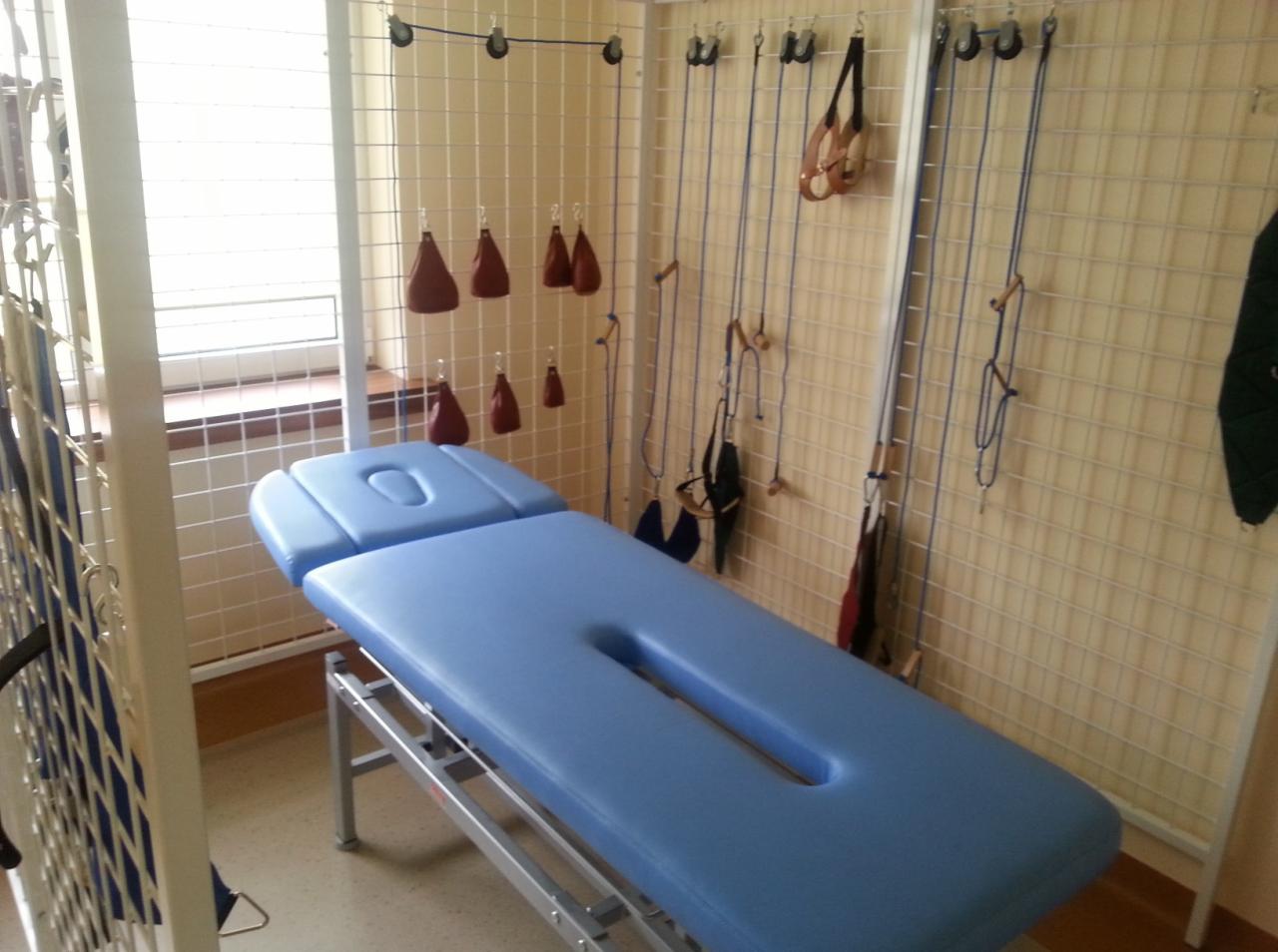Niebieski stół rehabilitacyjny z wgłębieniami, widoczne akcesoria do ćwiczeń wiszące na ścianie z kratką, jasne pomieszczenie z oknem.