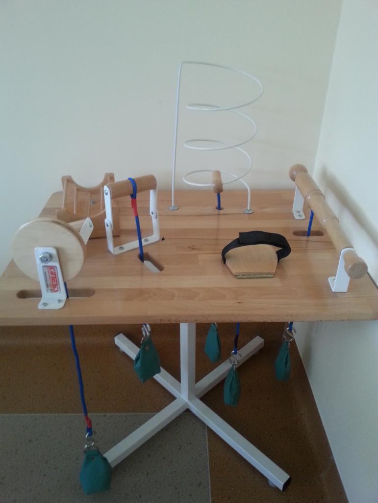 Stół rehabilitacyjny z różnorodnymi przyrządami do ćwiczeń manualnych, w tym labirynt, uchwyty i obciążniki. Sprzęt do terapii ręki.