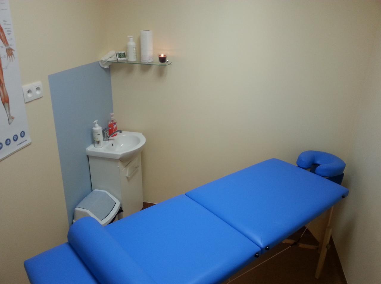 Niebieski stół do masażu z zagłówkiem w gabinecie. Umywalka z dozownikiem, półka ze świecą i schemat ciała na ścianie.