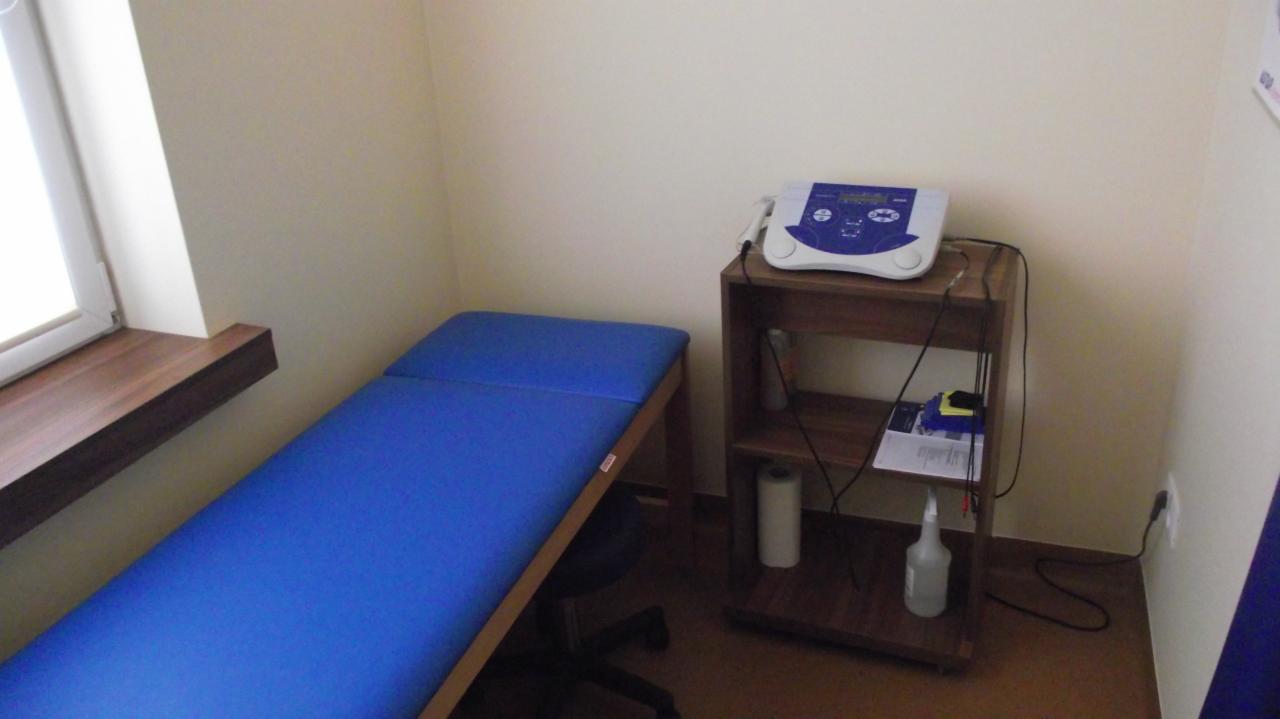 Niebieski stół do masażu w gabinecie z drewnianym regałem, na którym stoi urządzenie do elektroterapii. Przy oknie drewniany parapet.