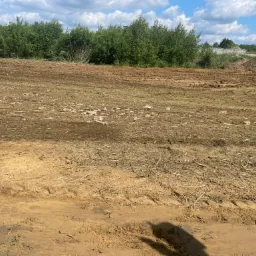 Przygotowany teren pod budowę z widocznymi śladami opon, usuniętą roślinnością i wyrównaną ziemią w słoneczny dzień.