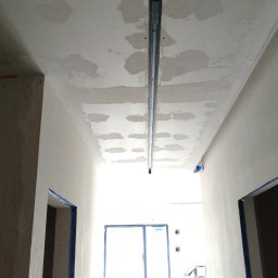 NewLifeColor MAREK BECHT Malowanie Mieszkań i Domów - Sufit w trakcie montażu profili, widoczne ślady szpachlowania. W tle drzwi z niebieską ramą i oświetlenie studyjne. Ujęcie z dołu, perspektywa korytarza.