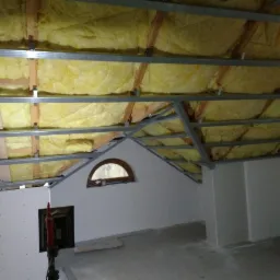 Poddasze w trakcie adaptacji: widoczna izolacja z wełny mineralnej między krokwiami, metalowy stelaż pod płyty gipsowo-kartonowe oraz fragment ściany z małym, półokrągłym oknem.