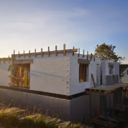 Wczesny etap budowy domu z białych bloczków, widoczne szalunki okienne i drewniane podpory, fundament zabezpieczony izolacją, w tle błękitne niebo i zieleń drzew.