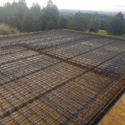 Zbrojenie płyty fundamentowej z widocznymi elementami dystansowymi z tworzywa, przygotowane do zalania betonem, na tle zalesionego terenu i zabudowy miejskiej w oddali.