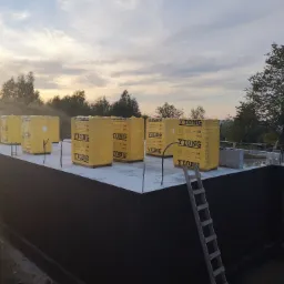 Fundamenty domu z izolacją pionową, przygotowane do murowania ścian z bloczków YTONG, widoczna drabina oparta o ścianę fundamentową, w tle drzewa i zachodzące słońce.