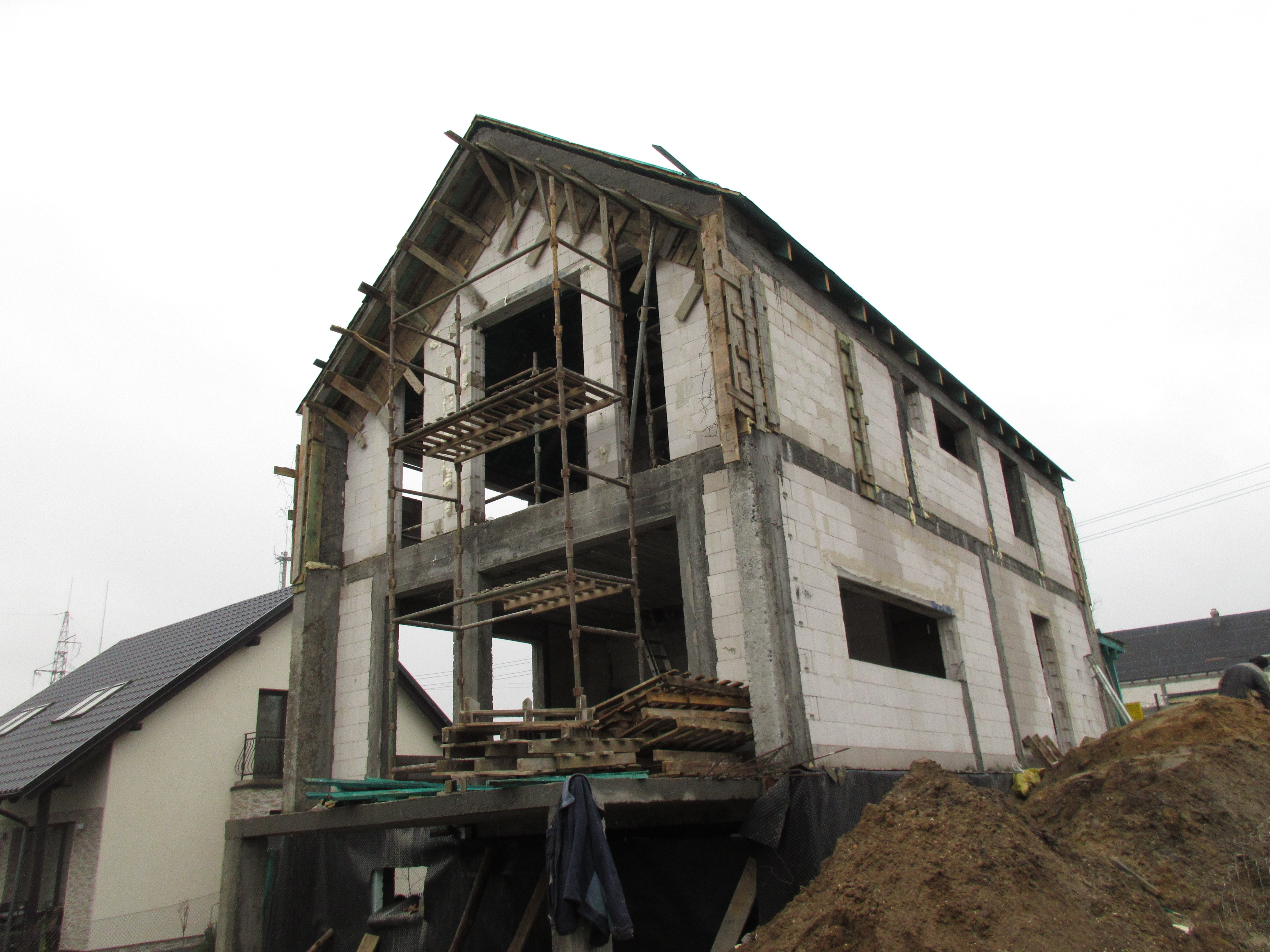 Surowy stan domu z białych bloczków, w trakcie budowy, widoczne rusztowania i drewniane elementy konstrukcyjne, z ziemią usypaną przy ścianie budynku.