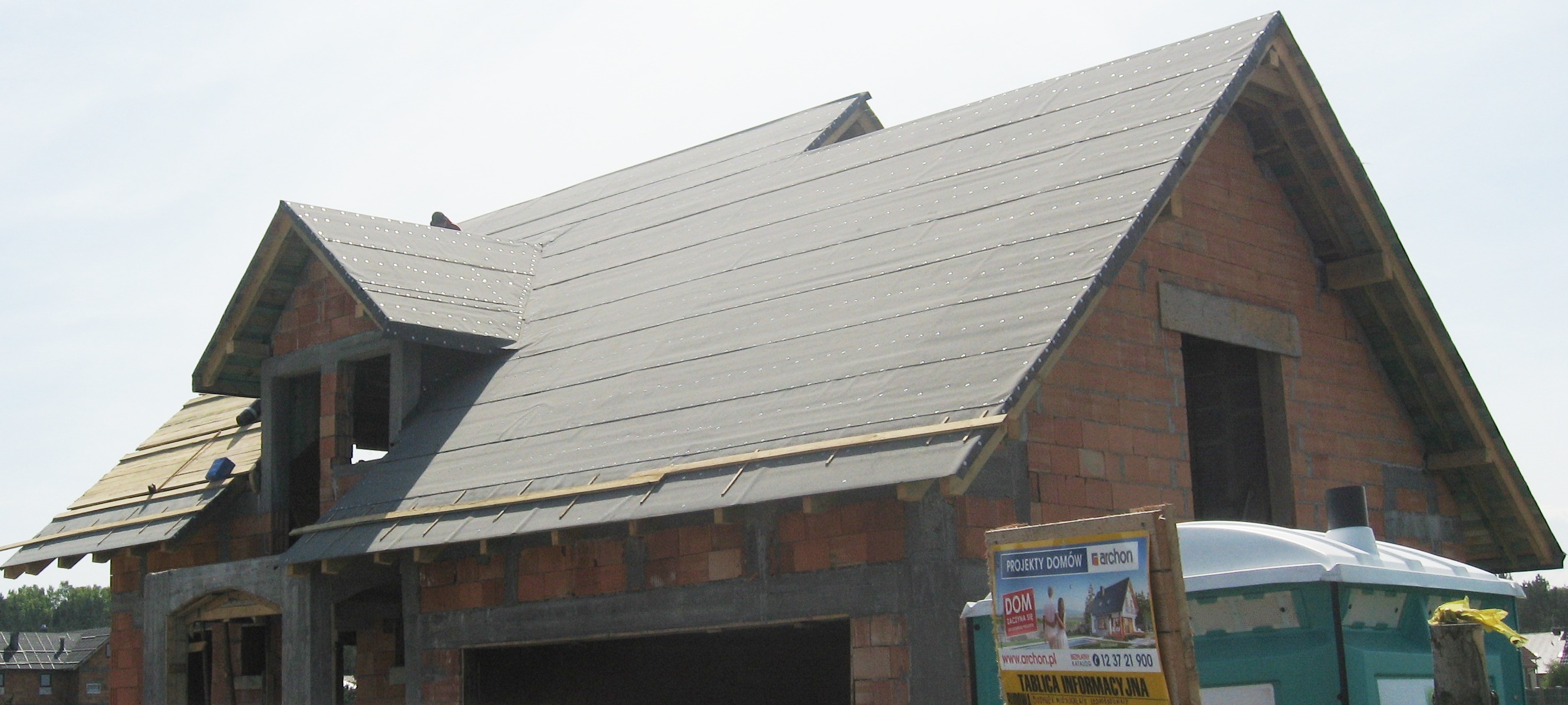 Dom murowany w trakcie budowy, widoczna konstrukcja dachu z membraną, ściany z czerwonej cegły, otwarte okna, tablica informacyjna budowy i przenośna toaleta.